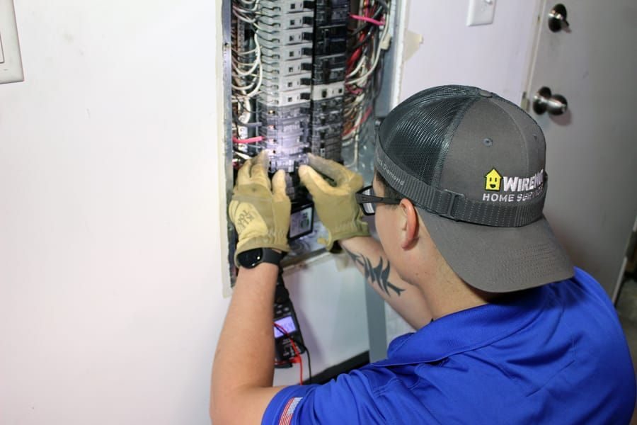 Electrical technician in a blue shirt and gloves adjusting breakers inside a wall-mounted electrical panel.