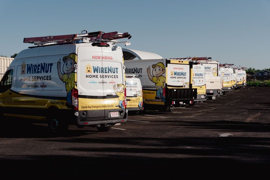 Line of WireNUT Home Services vans with ladders on top, parked in a lot; 'Now Hiring' signs visible on the backs of some vehicles.
