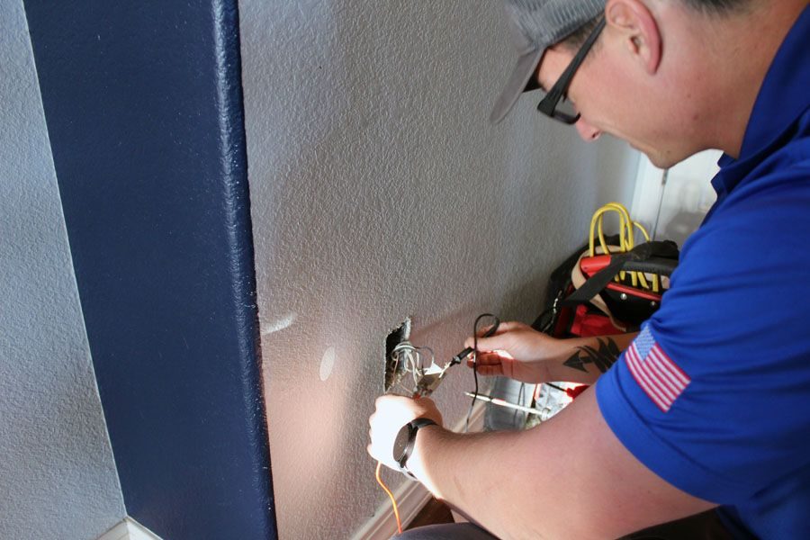 Electrician in safety glasses repairs exposed wiring in a wall outlet at a baseboard.