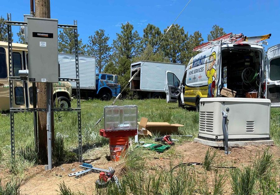 Outdoor service site with a Generac electrical panel mounted on a metal frame, a large generator to the right, and a yellow service van with ladder graphics in the background.