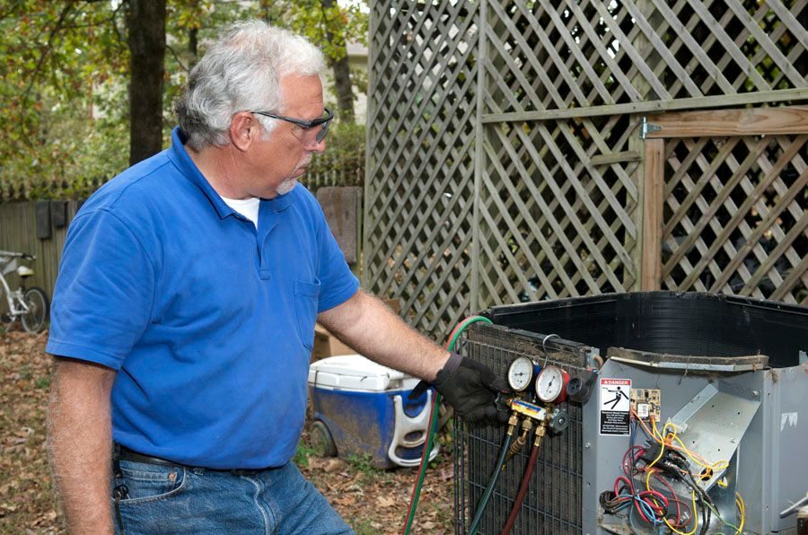 Man in a blue polo shirt and gloves checks refrigerant gauges on an outdoor air conditioning condenser unit in a yard.