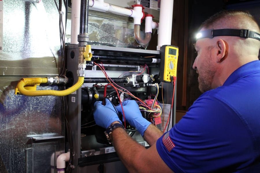 HVAC technician in gloves and a headlamp works on an exposed furnace control panel with wires and tubing nearby.