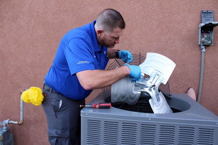 HVAC technician in a blue shirt and gloves removing a metal component from an outdoor condenser unit with a screwdriver nearby on top of the unit.