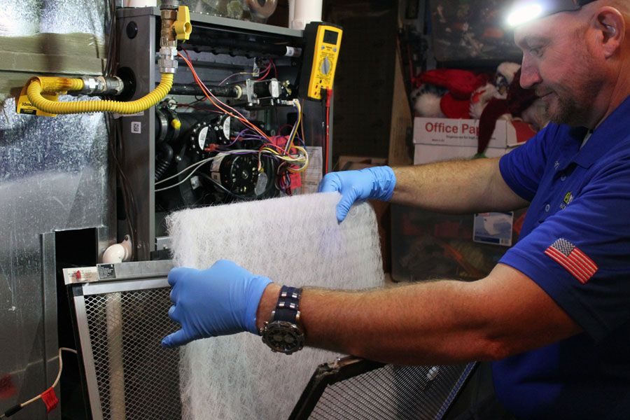 HVAC technician wearing blue gloves and a blue shirt removes a white air filter from a furnace or air handler unit.