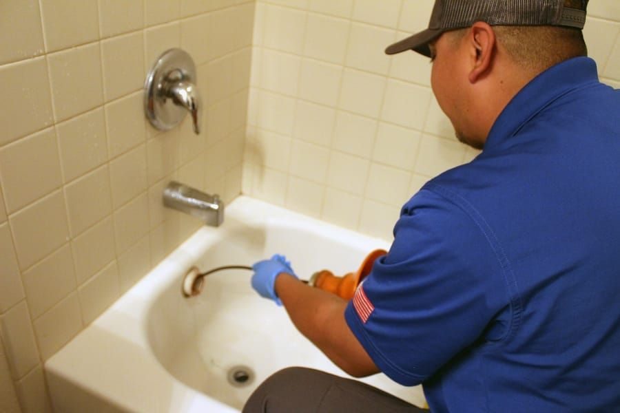 Plumber in a blue uniform using a tool to clear a bathtub drain, wearing gloves and a cap in a bathroom.