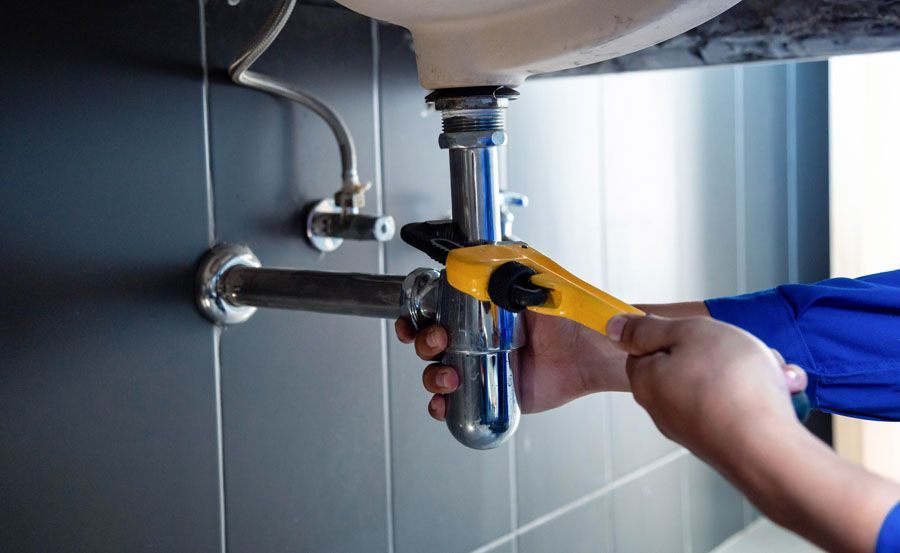 Person in a blue sleeve uses a yellow adjustable wrench to tighten a chrome plumbing pipe under a sink.