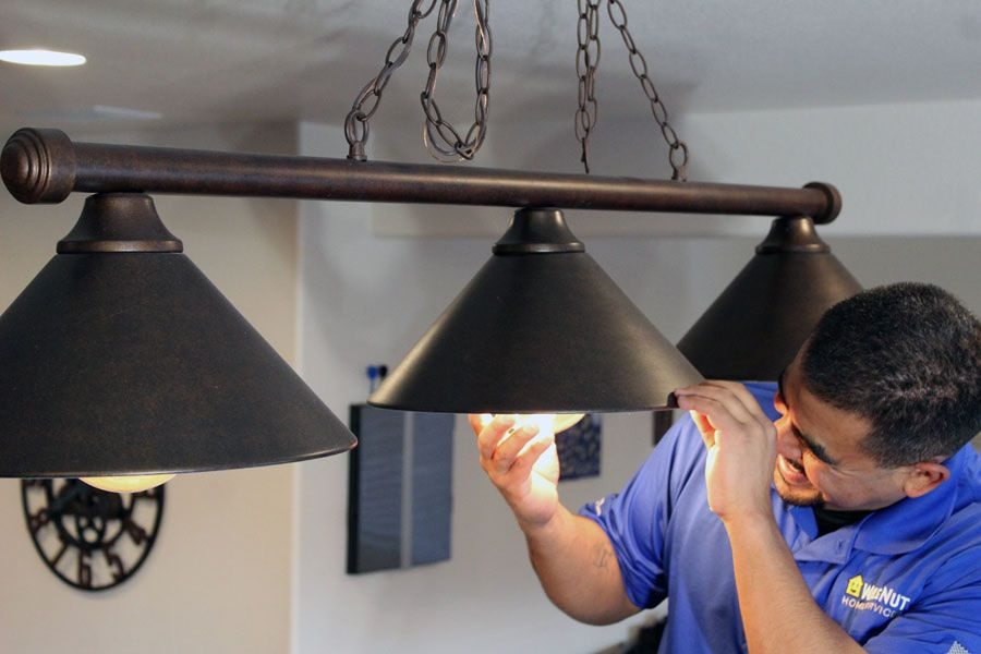 Technician in a blue shirt adjusts a row of black pendant lights hanging from the ceiling, chains visible above. — Indoor lighting installation scene