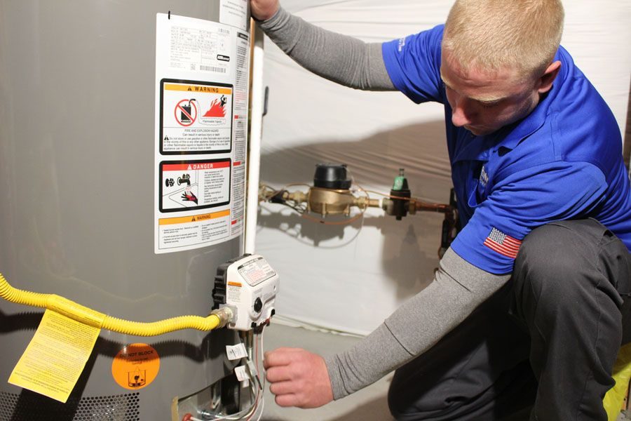 Technician in a blue shirt servicing a water heater, kneeling and adjusting a valve with a yellow gas line nearby (warning labels visible).