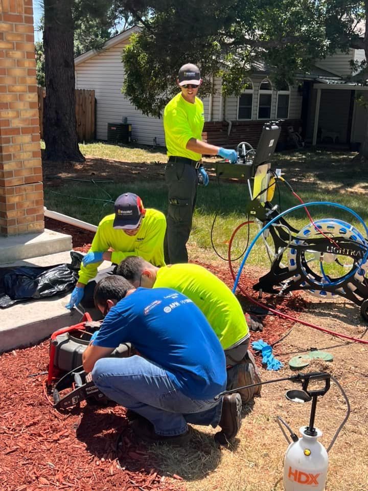 Three workers in neon shirts perform outdoor utility work by a house; one stands at a laptop on a cart while two crouch and handle equipment and cables.